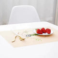 A beige rectangular placemat on a white table, styled with a gold and white fork, a small white plate with three red strawberries, and a white chair in the background.